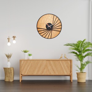 May include: A wooden wall clock with black metal accents and a sunburst design. The clock is hanging on a white wall above a wooden dresser with a light wood finish.