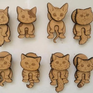 May include: A set of eight light brown wooden cat-shaped drawer knobs, each featuring a unique laser-engraved feline design with distinct fur patterns, facial expressions, and tail shapes, arranged in two rows of four on a plain surface.