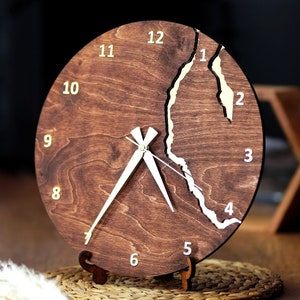 Puede incluir: Un reloj de madera marrón con manecillas blancas y números dorados. La esfera del reloj tiene un diseño de grieta blanca. El reloj está sobre un soporte de madera.