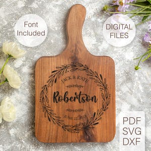 May include: A wooden cutting board with a handle, engraved with a wreath design and the text "Jack & Kate Robertson June 22, 2023".