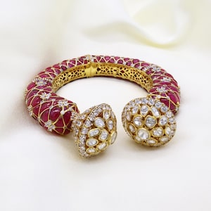Puede incluir: Un brazalete dorado y pendientes a juego. El brazalete presenta piedras preciosas rojas y pequeños diamantes, con un diseño de filigrana. Los pendientes son redondos, con un grupo de gemas claras. La joyería se encuentra sobre un fondo blanco.