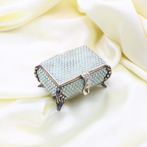 May include: A small, rectangular jewelry box with a light blue, scale-like pattern on the lid and sides. The box has silver-colored metal trim, feet, and a clasp. The box is set against a backdrop of a cream-colored, silky fabric.