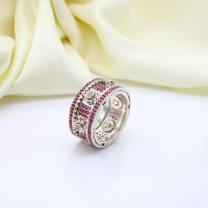 Puede incluir: Un anillo plateado con una banda de pequeñas piedras preciosas rojas redondas y piedras preciosas claras redondas. El anillo tiene un diseño detallado con un acabado pulido. El fondo es una tela suave de color blanquecino.