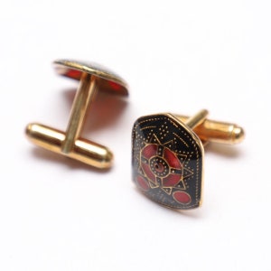 Puede incluir: Un par de gemelos de oro con diseños de esmalte negro y rojo. Un gemelo presenta un patrón geométrico con un centro rojo, mientras que el otro gemelo tiene un diseño más simple.