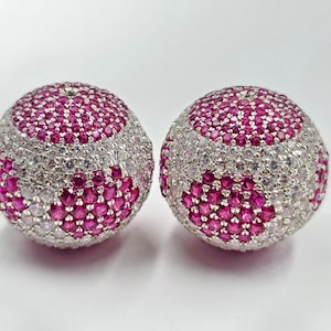 Könnte beinhalten: Zwei runde Perlen mit einem rosa-weißen Design. Die Perlen sind mit kleinen rosa und weißen Edelsteinen besetzt.