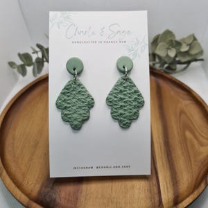 Puede incluir: Un par de pendientes verdes de declaración con un diseño de hoja texturizada. Los pendientes están hechos a mano y presentan un pequeño encanto redondo verde en la parte superior.