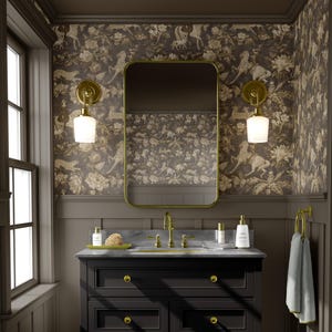 May include: A bathroom vanity with a black cabinet, gold fixtures, and a marble countertop. The mirror has a gold frame. The wallpaper features a floral and animal pattern in shades of brown and beige. Two sconces with white glass shades flank the mirror.
