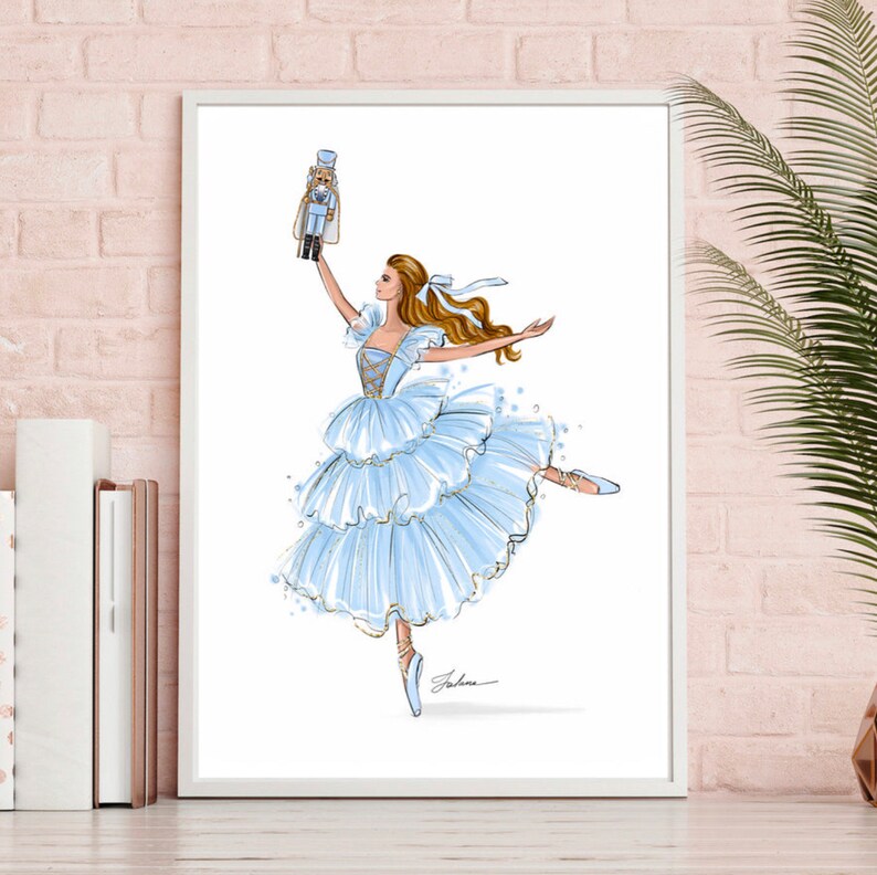 May include: A ballerina in a blue and gold tutu holds a nutcracker doll above her head. She is dancing on pointe with her leg extended. The background is white.