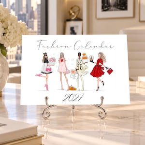 Pode incluir: Um cartão retangular branco Fashion Calendar 2027 com quatro figuras ilustradas em trajes elegantes, incluindo um vestido preto e branco, um vestido rosa, um terno bege e um vestido vermelho, exibido num suporte de metal prateado.