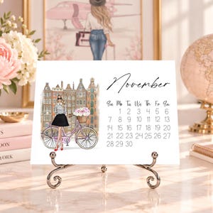 Pode incluir: Um cart&atilde;o de calend&aacute;rio de novembro num suporte de metal com a ilustra&ccedil;&atilde;o de uma pessoa com camisa &agrave;s riscas e saia preta ao lado de uma bicicleta cor-de-rosa com um cesto de flores em frente a edif&iacute;cios europeus altos e estreitos.