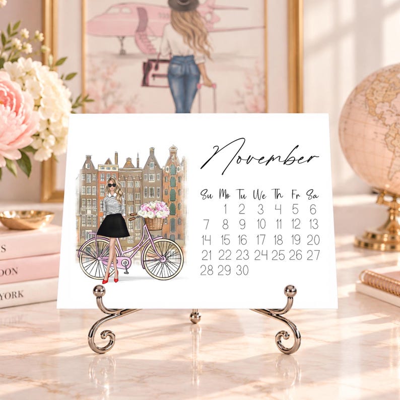 Pode incluir: Um cart&atilde;o de calend&aacute;rio de novembro num suporte de metal com a ilustra&ccedil;&atilde;o de uma pessoa com camisa &agrave;s riscas e saia preta ao lado de uma bicicleta cor-de-rosa com um cesto de flores em frente a edif&iacute;cios europeus altos e estreitos.