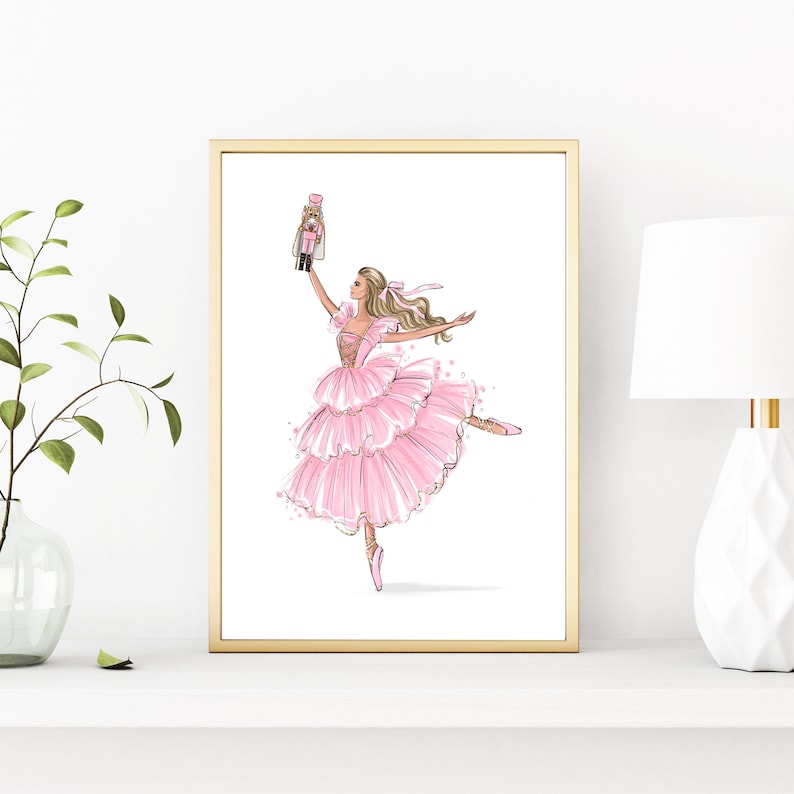May include: A framed illustration of a ballerina in a pink tutu holding a nutcracker doll. The ballerina is wearing pink ballet shoes and has blonde hair. The background is white.
