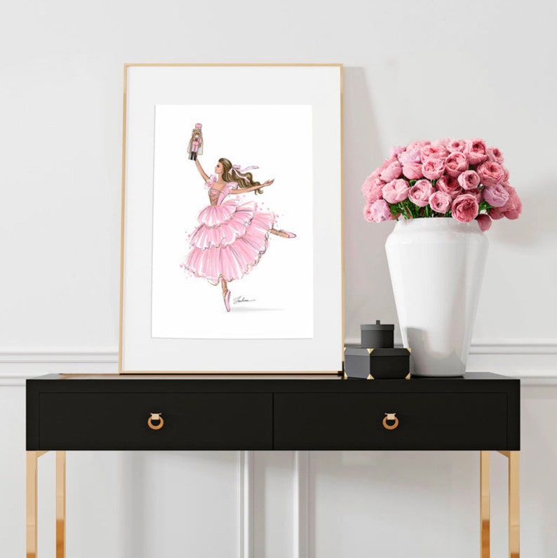 May include: A framed illustration of a ballerina in a pink tutu holding a nutcracker doll. The ballerina is dancing with her leg extended and her arms raised. The background is white.