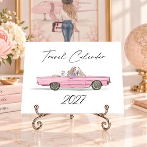 Pode incluir: Um calendário de viagem 2027 branco num suporte de metal com uma ilustração de um carro descapotável cor-de-rosa com uma pessoa e um cão branco pequeno lá dentro. O texto Travel Calendar está em preto acima e 2027 abaixo.