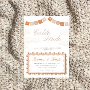 Op de afbeelding: Een baby shower uitnodiging met een lichtblauwe achtergrond en oranje en witte decoraties. De tekst luidt "Cielito Lindo" en "Bianca & Dario". De uitnodiging bevat de datum, tijd, locatie, RSVP-informatie en registratie-informatie.