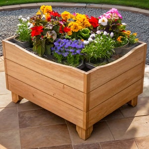Può includere: Un vaso da fiori in legno con più scomparti, pieno di fiori colorati, tra cui fiori rossi, gialli, bianchi e viola. Il vaso è rialzato su quattro gambe e si trova su un patio in pietra.