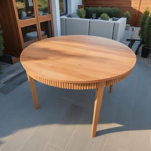 Puede incluir: Una mesa redonda de madera con un borde acanalado y cuatro patas. La mesa es de madera marrón claro y está sobre un patio de baldosas grises.