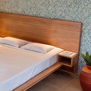 Puede incluir: Un marco de cama de madera con una cabecera y una colcha blanca. La cama tiene dos mesitas de noche con estantes. Una planta en maceta se encuentra en una maceta de terracota en el suelo.