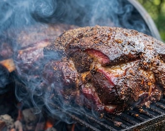 May include: A large, dark brown, seasoned pork shoulder roast cooking on a black metal grill grate. The meat has a textured, crusted surface with visible spices and is surrounded by thick, swirling white smoke rising from the hot coals below.