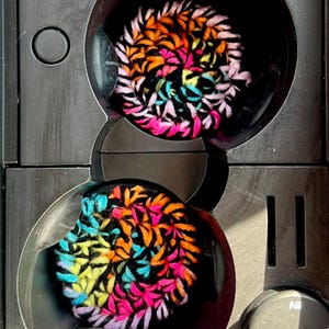 May include: Two colorful crocheted coasters in a black holder. The coasters are made with a variety of colors, including pink, orange, blue, and yellow. The holder has two circular openings and a small black button with the text "5V" on it.