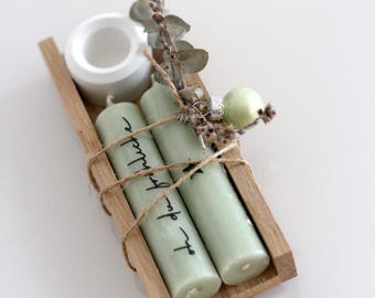 May include: A wooden tray displays two pale green candles with black script, bound with twine. A white ceramic candle holder, dried greenery, and a small green bauble are also present. The candles are approximately 15 cm tall.