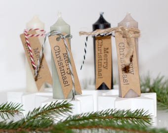 May include: Four taper candles in white, sage green, black, and taupe, each set in a white star-shaped base. Every candle features a brown paper tag tied with string, printed with the text Merry Christmas! in black ink.