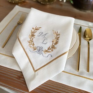 May include: White linen napkin with gold embroidered initials inside a laurel wreath. The napkin has a gold border and is folded on a white plate. Gold silverware is arranged on a white placemat.