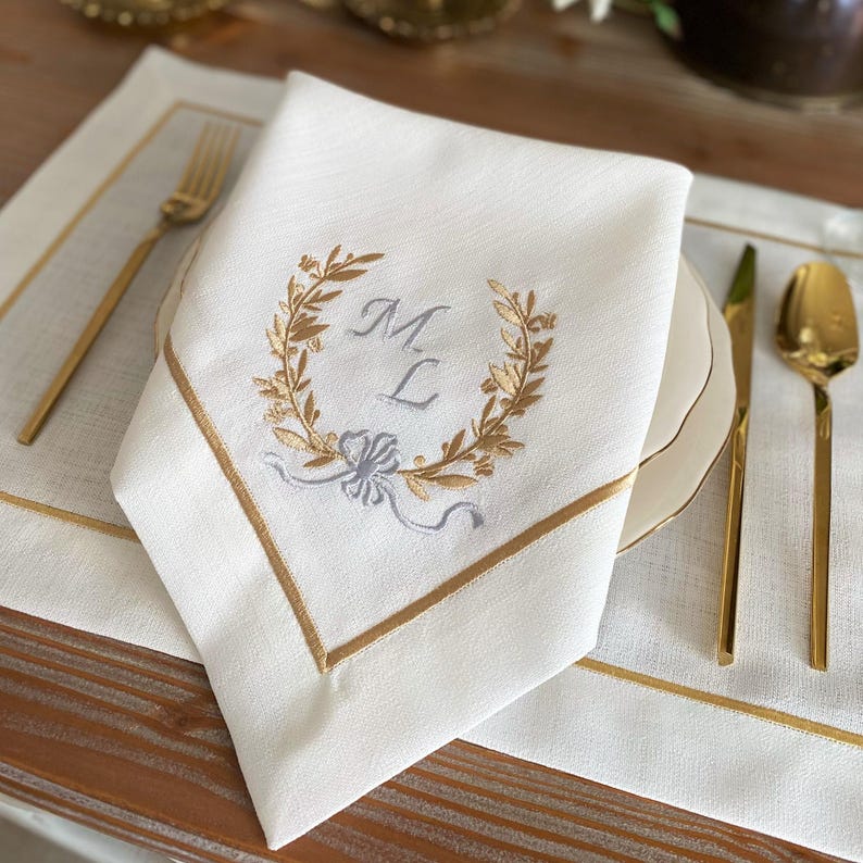 May include: White linen napkin with gold embroidered initials inside a laurel wreath. The napkin has a gold border and is folded on a white plate. Gold silverware is arranged on a white placemat.