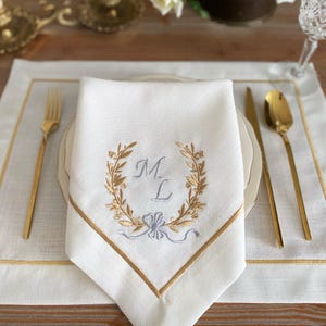 Könnte beinhalten: Eine weiße Leinen-Serviette mit goldenem Rand und gestickten Initialen in einem goldenen Lorbeerkranz. Die Serviette liegt auf einem passenden Tischset, zusammen mit goldenem Besteck, wodurch eine elegante Tischdekoration entsteht.
