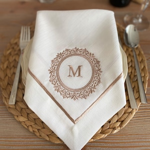 May include: White linen napkin with an embroidered border and the letter "M" in a circular design. The napkin is folded and placed on a plate with a fork and spoon on a woven placemat.