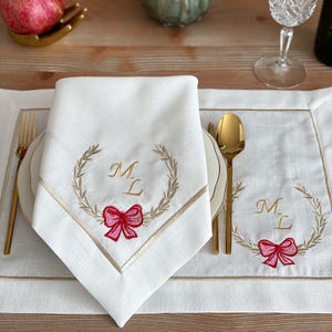 Peut inclure: Ensemble de nappe et de nappe en lin blanc avec des bordures dorées et des détails brodés. La serviette présente un motif de couronne avec les initiales "M L" et un nœud rose. Une fourchette et une cuillère dorées sont sur la table.