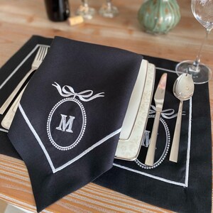 Peut inclure: Une serviette de table noire avec un motif ovale brodé blanc avec la lettre "M" et un nœud. La serviette fait partie d'une mise en place avec des couverts et une assiette décorative.