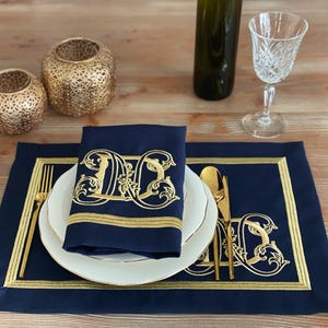 May include: A navy blue placemat and napkin set with gold embroidered details. The napkin features a gold monogram design. Gold-colored silverware and a wine glass are also present, creating an elegant table setting.