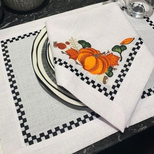 May include: White linen napkin featuring an embroidered orange pumpkin and autumn leaves. The napkin rests on a black and white striped plate, placed on a placemat with a black chequered border. A seasonal table setting.