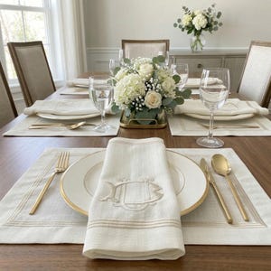 Peut inclure: Une table à manger joliment dressée avec des assiettes blanches, des couverts dorés et des serviettes en lin blanches. Un centre de table floral de roses et d'hortensias blancs est dans un récipient doré. Verres à eau et à vin.