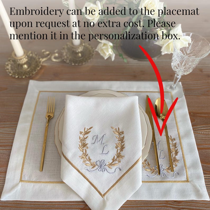May include: A white linen placemat and matching napkin set with gold embroidery. The napkin features a monogram within a laurel wreath design. Gold-colored flatware and a decorative border complete the elegant table setting.
