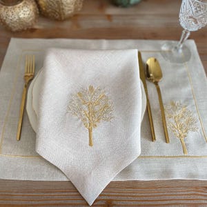 May include: A table setting with a cream-colored linen napkin featuring a gold embroidered tree design. The napkin is placed on a white plate, with gold cutlery and a matching placemat. A crystal wine glass is visible.