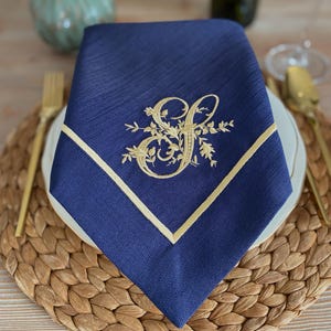 May include: A navy blue napkin with a gold embroidered initial "S" and floral detailing. The napkin has a gold border and is on a white plate with gold cutlery. The napkin is placed on a woven placemat.