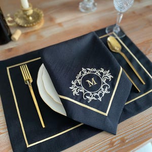 Könnte beinhalten: Eine schwarze Leinen-Serviette mit goldenem Rand und einem gestickten Monogramm "M" in einem dekorativen Rahmen. Die Serviette liegt auf einem passenden Platzset, mit goldenem Besteck und einem weißen Teller. Das Arrangement befindet sich auf einem Holztisch.