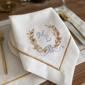 May include: White linen napkin with gold embroidered trim and a gold and gray embroidered wreath design. The wreath surrounds the gray embroidered initials "M L". A light blue bow is embroidered below the initials.
