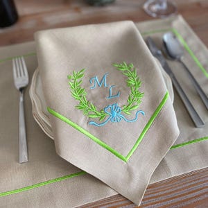 Peut inclure: Serviette en lin beige avec des détails brodés verts et bleus. Comprend une couronne de laurier verte avec les initiales "ML" en bleu et une bordure verte assortie. La serviette est posée sur une assiette blanche, avec une fourchette et une cuillère visibles.
