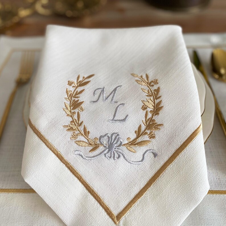 May include: White linen napkin with gold embroidered trim and a gold and silver embroidered wreath design. The wreath surrounds the letters "M L". The napkin is folded and placed on a white placemat with gold trim.