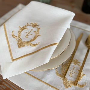 Könnte beinhalten: Weiße Leinen-Serviette mit goldgestickten Initialen und dekorativem Rand. Die Serviette ist gefaltet und auf einem weißen Teller platziert, mit goldenem Besteck und einem passenden Tischset.