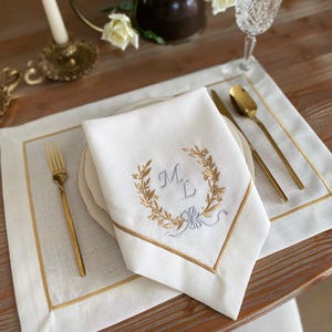 May include: White linen napkin with gold embroidered laurel wreath and initials, set on a matching placemat. Gold trim borders the napkin and placemat. Gold fork, knife, and spoon complete the elegant table setting.