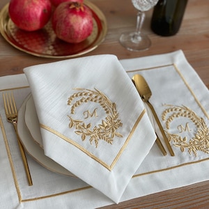 Könnte beinhalten: Weißes Leinen-Servietten- und Platzset mit goldgesticktem Blumenkranz und dem Buchstaben "M". Goldbesatz umrandet die Serviette und den Tischset. Goldene Gabel und Löffel liegen auf dem Tisch. Granatäpfel liegen auf einem goldenen Tablett.