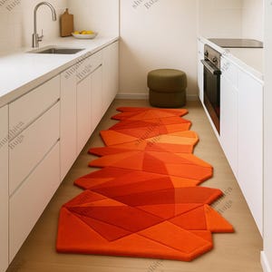 Puede incluir: Una alfombra de pasillo geométrica naranja con un diseño moderno. La alfombra presenta una serie de formas angulares interconectadas en varios tonos de naranja, creando un efecto visual llamativo. La alfombra está colocada en una cocina.