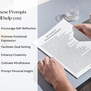 May include: A person holding a white paper with "100 Self-Reflection Journaling Prompts" printed on it. The paper is on a gray surface. Text on the paper includes prompts to encourage self-reflection, promote emotional expression, and facilitate goal setting.