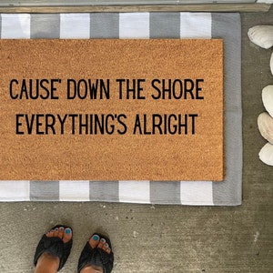 May include: A brown coir doormat with the text "CAUSE' DOWN THE SHORE EVERYTHING'S ALLRIGHT" in black lettering. The doormat is on a gray and white striped rug.