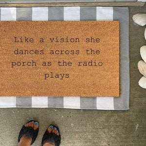 May include: A rectangular doormat with the text "Like a vision she dances across the porch as the radio plays." The mat is brown and sits on a gray and white striped rug. White seashells are scattered nearby.