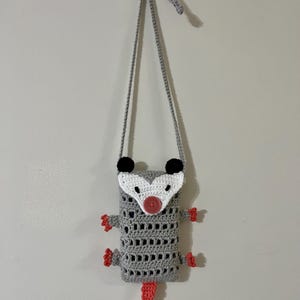 May include: A handmade crochet possum bag in gray with a white face, black ears, and a pink nose and tail. The bag has a gray strap and red feet. The bag is hanging on a white wall.